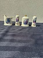 Full set of four pieces - creamer, sugar jar, salt and pepper shakers from Thomas Kinkade Sleeping Cottage Avon collection on a dark background against a light wall.