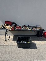 Full view of all the tools laid out on a black folding table outdoors against a wall and a black bin in front containing more tools.