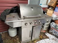 Front view of the stainless steel Terra Gear propane BBQ with four control knobs, side burner on left, two propane tanks under cabinet, double-door storage cabinet, positioned next to a red wall with leaves and snow on the ground, showing weathering and dirt.