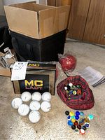 View of boxed MC Lady golf balls, loose white golf balls, marbles in a red pouch and loose on floor, and part of the bocce ball set