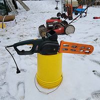 Front-side view of electric chain saw with orange blade guard, sitting on a yellow bucket in a snowy yard, showing partial lawn mower and other equipment in the background.