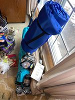 Photo showing rolled collapsible cot in blue carrying case, blue inflatable ground sheet, lanterns, flashlights, radio, and binoculars packed together on floor.