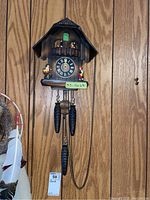 Full view of the wooden cuckoo clock hanging on a wall, showing the chalet style, clock face, figurines, and pinecone weights.