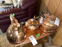 Photo showing the three copper kettles side by side on a wooden bench with visible dents and marks, with one kettle exhibiting a wood spindle-style handle.