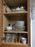 Cabinet shelf with stacked Noritake Majestic Gold dishes, including plates, cups, saucers, and two gold salt and pepper shakers
