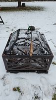 Front angled view of square metal fire pit with mesh sides and closed mesh top, poker tool resting on top, with snow on the ground.