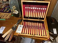 Photo of the open wooden box with 12 fish knives and 12 fish forks arranged inside, showing cream faux ivory handles and decorative metal tops on a red velvet and satin lining.
