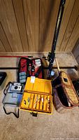 Full view of lot including two fishing rods with reels, yellow tackle box open with lures visible, wicker fishing basket, fishing net, and Polaroid Land Camera with case on carpet.