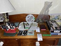 Full lot overview on a wooden dresser showing ceramic figurines, manicure sets, jewelry pieces, wristwatches, decorative plate, boxed domino trays, and a New Testament book.