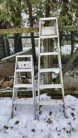 Two aluminum step ladders standing next to each other outside on snow with a chain link fence background. One ladder is smaller (4 feet) and the other taller (6 feet).