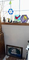 Photo showing stained glass sun catchers on a window ledge, including hexagonal designs, a square bird piece, a butterfly figure, framed loon artwork below, and figurines including a dragon, cat, and penguins.