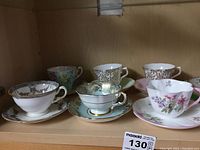 Six assorted decorative teacups and saucers on wooden shelf, showcasing unique patterns and colors