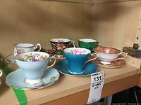Six sets of teacups and matching saucers on wooden shelf, showing variety of floral patterns and colors.