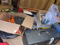 Cardboard boxes with hardware items and coiled rope beside metal toolbox.