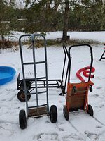 Three moving dollies placed outdoors on snow-covered ground, showing two metal hand trucks and one wooden base hand truck.