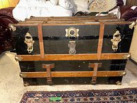 Front view of vintage High Carson trunk showing black exterior, wood slats, leather straps, brass clasps and corner protectors.