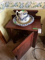 Full view of the wooden washstand with open storage door and blue and white ceramic pitcher and basin placed on top, showing towel racks on each side and carved backboard.
