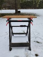 Front view of collapsible work bench showing wooden top panels and metal folding frame in snowy outdoor setting.