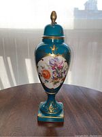 Front view of the Limoges urn showing teal blue background, gold accents, and colorful floral painting on porcelain surface with finial on top