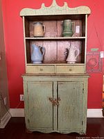 Full front view of the antique cupboard showing the green painted finish with visible crackling and red underlayer, open shelf area with ceramic pitchers, two small drawers with round knobs under the open shelf, and the two-door lower cabinet secured with leather strap lock.