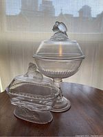 Full side view of both pressed glass items on table, showing the comport with doves on lid and rectangular covered dish with lion on lid and base.
