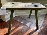 Side and angled view of antique primitive wooden footstool showing its rectangular top and four angled legs on wooden floor.