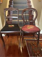 Front view of two antique wooden chairs with floral embroidered needlepoint seats placed on wooden floor in front of stairs.