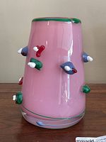 Front view of pink cylindrical art glass vase with applied multicolored glass decorations on the sides. Glossy finish with green rim.