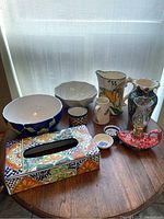 Overview of all ceramic items on wooden table including tissue box cover, bowls, vases, cups, jug, and oil lamp