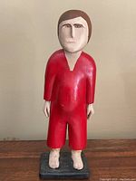 Full view of the hand-carved wooden sculpture depicting a man standing on a black base, wearing a red one-piece garment and barefoot with repaired ankles.