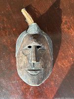 Front view of dark brown wooden mask with carved face and fiber top knot.