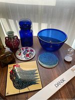 Nine items displayed on a wooden table include two glass vases, a blue glass bowl, a blue lidded container, two ceramic trivets, a small hourglass, and a boxed set of Dansk candles.