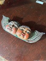 Wooden Mexican wall hanging with two carved faces and wings, showing front angle and corner repair.