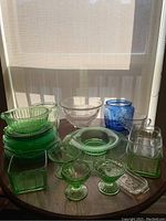 Photo of assorted green, clear, and blue Depression glass pieces including plates, bowls, cups, and butter dishes arranged on a small round table near window light.