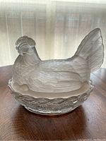 Photo showing frosted glass hen sitting atop the clear glass basket base on a wooden surface with light filtering through a curtain in the background.