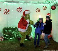 Photo of two children posing with a person dressed as the Grinch wearing a Santa costume, with festive peppermint decorations and green string lights in the background.