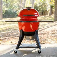 Red ceramic Kettle Joe grill on black wheeled stand with side shelves outdoors
