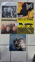 Front view of 5 vinyl record album covers by Queen, David Bowie, Steppenwolf, Men at Work, and Creedence Clearwater Revival on tiled floor.