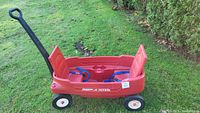 Side view of the red Radio Flyer kids wagon on grass, showing the handle, wheels, and two red folding seats with blue straps inside.