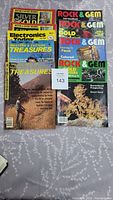 Photo showing a collection of eight magazines laid out on a gray patterned fabric. Magazines include Rock & Gem, Treasure, Silver & Gold, and Electronics Today International from the 1980s and 1990s.