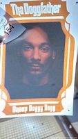 Close-up photo of a single iron on transfer featuring a portrait of Snoop Doggy Dogg with 'The Doggfather' text in orange and white.