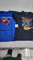 Two Spider-Man T-shirts laid flat; one royal blue with single Spider-Man graphic and one black with Spider-Man, spider web, and text graphic.