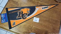 Photo of a vintage BC Lions CFL football pennant showing the team's roaring lion logo, BC letters, and CFL branding on an orange, black, and white fabric triangular pennant.