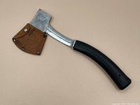 View of the hatchet with leather sheath covering the blade, showing curved black handle and metal axe head beneath the sheath.