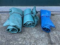 Three rolled tarps on concrete ground against a wall: two green woven tarps and one blue tarp, each tied with cords or elastic bands.