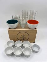 Photo showing two small patterned ceramic bowls with colored interiors on top of a wooden box, along with stacked small clear glass cups and several white ceramic ramekins below.