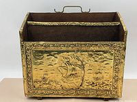 Front on photo showing the brass panel with embossed three sailing ships and surrounding floral pattern border on the magazine stand.