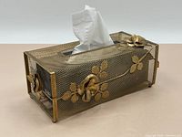 Showing the tissue box cover from an angled side view with tissue partially pulled through the top opening, displaying the metal mesh construction and attached metal flowers.