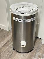 Front view of the stainless steel cylinder-shaped portable washing machine showing the water drainage spout and SpinX brand label.