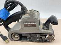 Close-up of Porter Cable 3x21 belt sander showing front and side, power cord coiled with blue tape, moderate dust and wear.